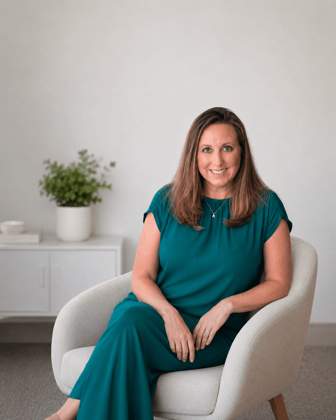 Smiling woman with long brown hair in a teal dress sits in a white armchair.