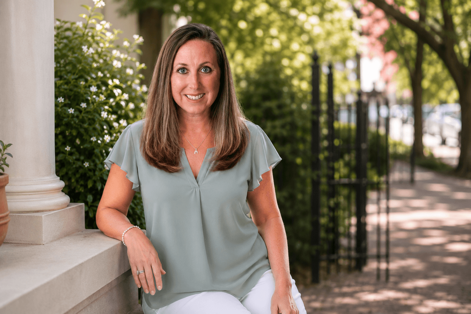Smiling woman in a sage green blouse leaning against a white pillar outdoors.